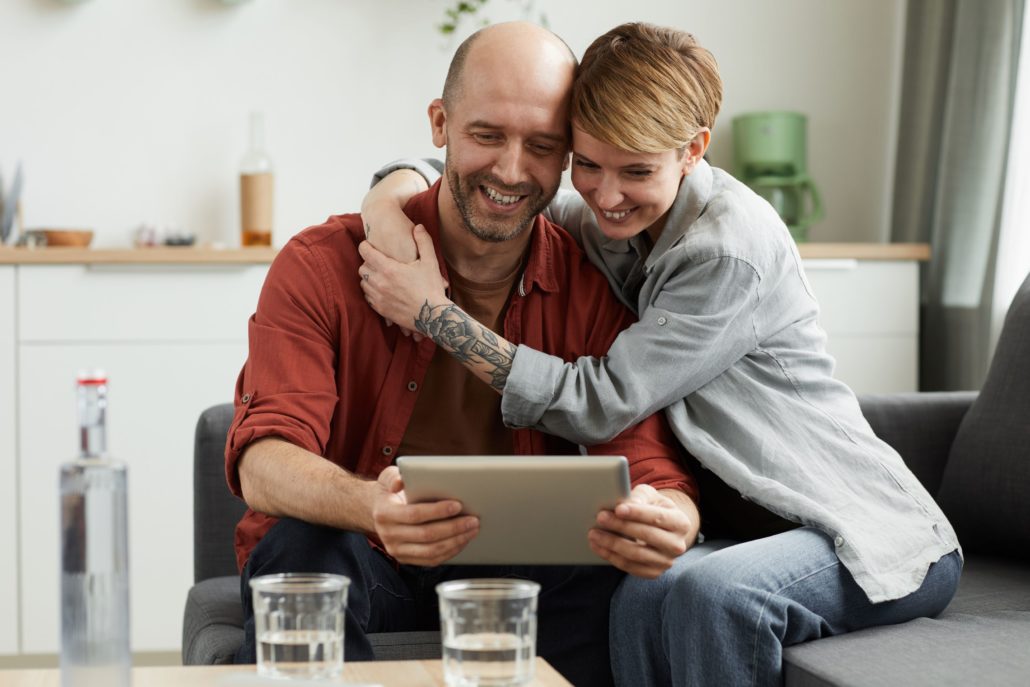 young smiling couple sitting o sofa and looking at digital tablet they talking to their friends online at home 1030x687